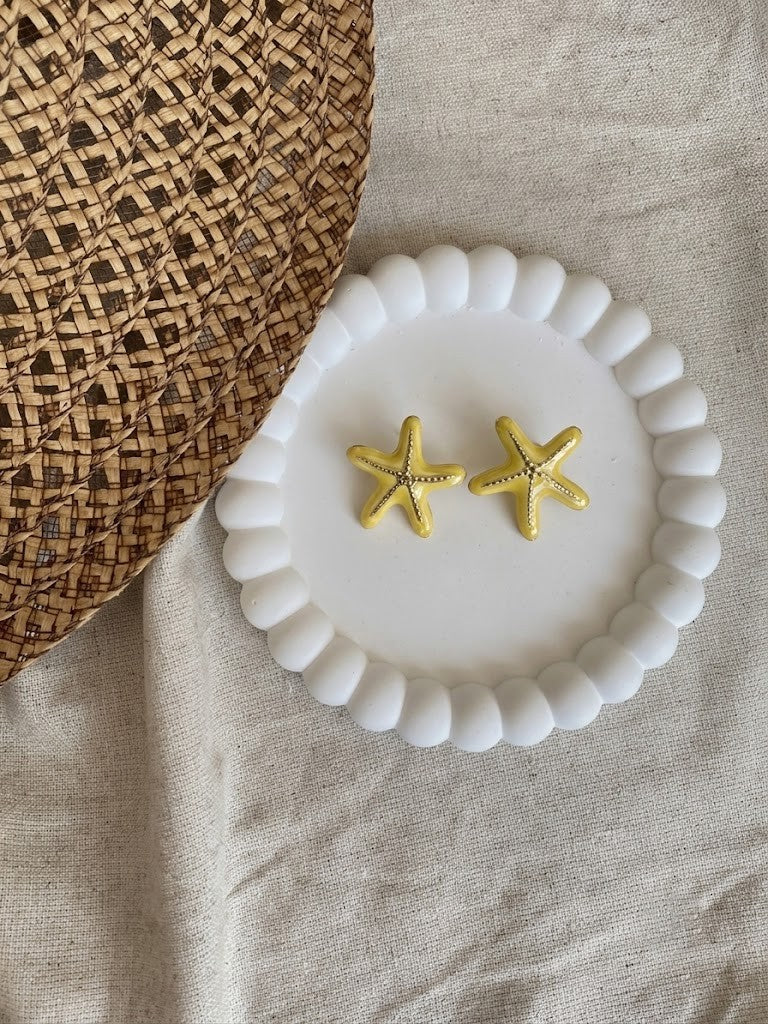 Gold starfish earrings on a white beaded dish with a woven mat and beige fabric background