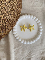 Gold starfish earrings on a white beaded dish with a woven mat and beige fabric background